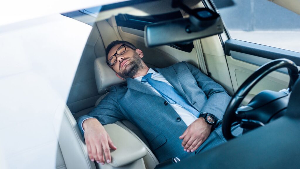 a man in a business suit sleeping in the driver's seat of his car.