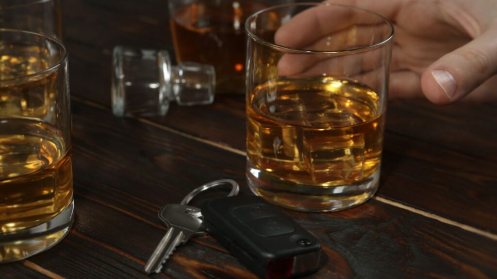 A man reaching for a glass of liquor with his car keys next to it.