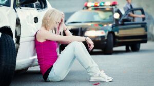 A young girl who is sitting next to her damaged car after a car accident.